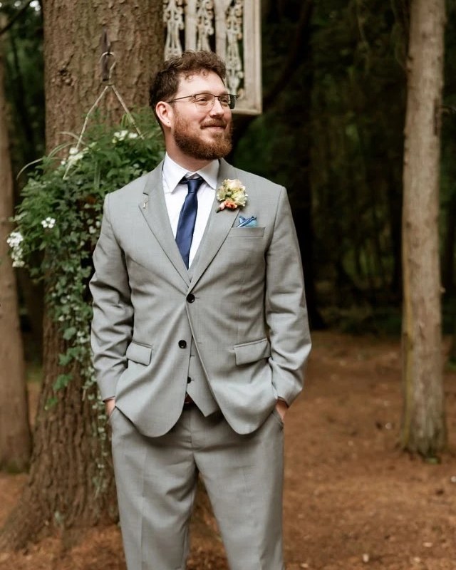 Dominic in a grey suit and blue tie, smiling and looking off to the side in a wooded clearing